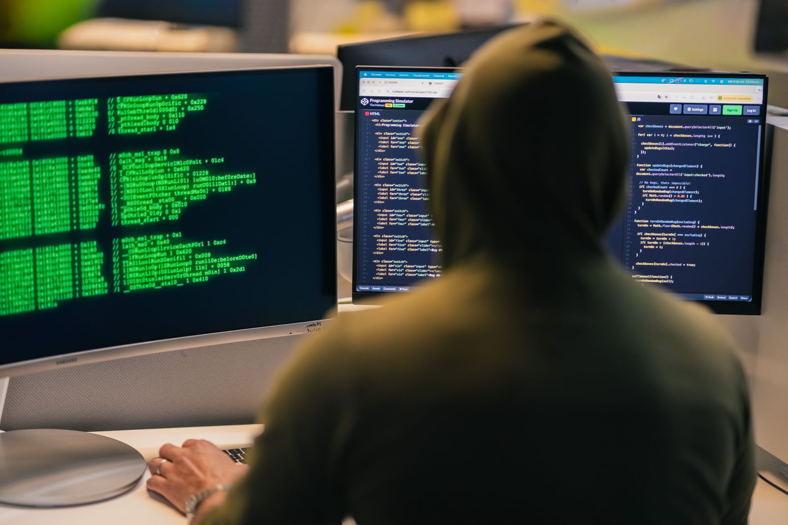 Hooded person working at two monitors filled with green code